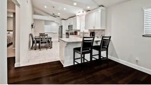 A modern kitchen with white cabinetry, a central island with chairs, and a dining area with a table and chairs. - Lake View! Hot-Tub! Beaver Lake House! (Rogers)