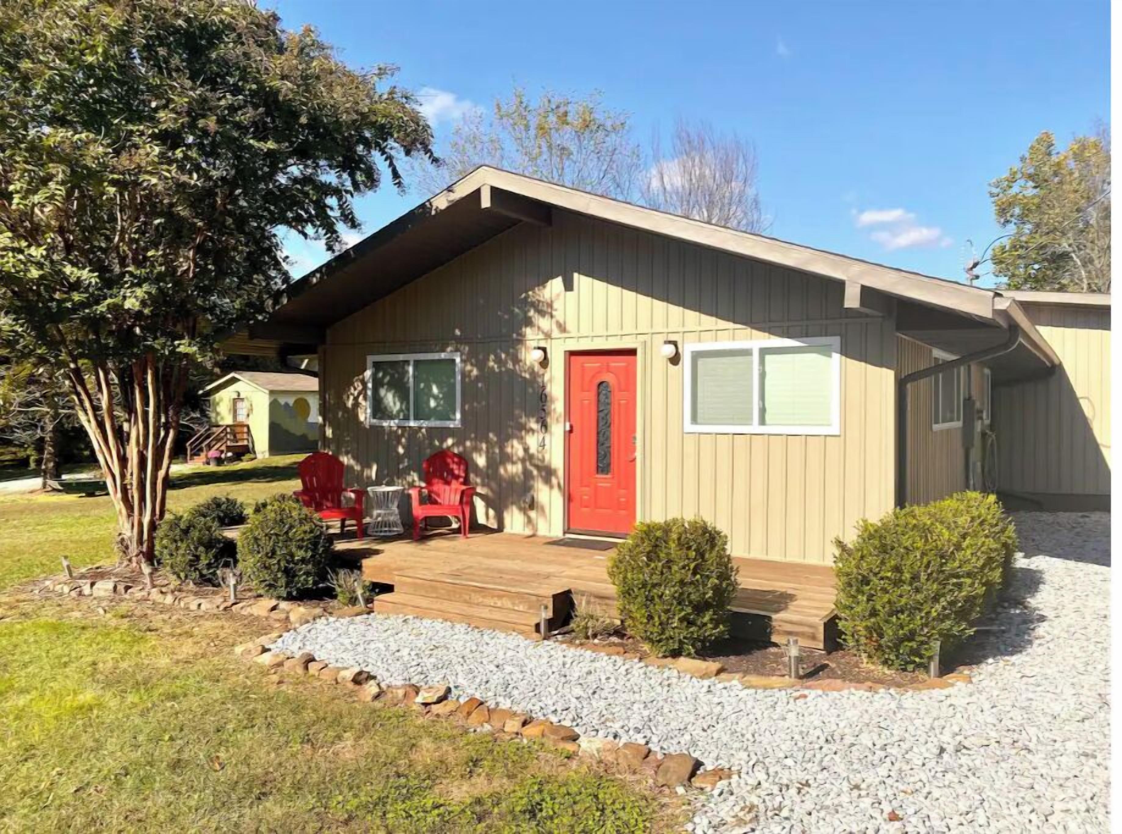 A single-story building with a red door, a covered porch, and a small garden area with gravel and plants.