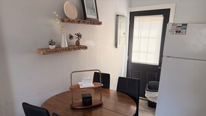 A dining area with a round wooden table, black chairs, a small wooden side table with a sign, and a white refrigerator.