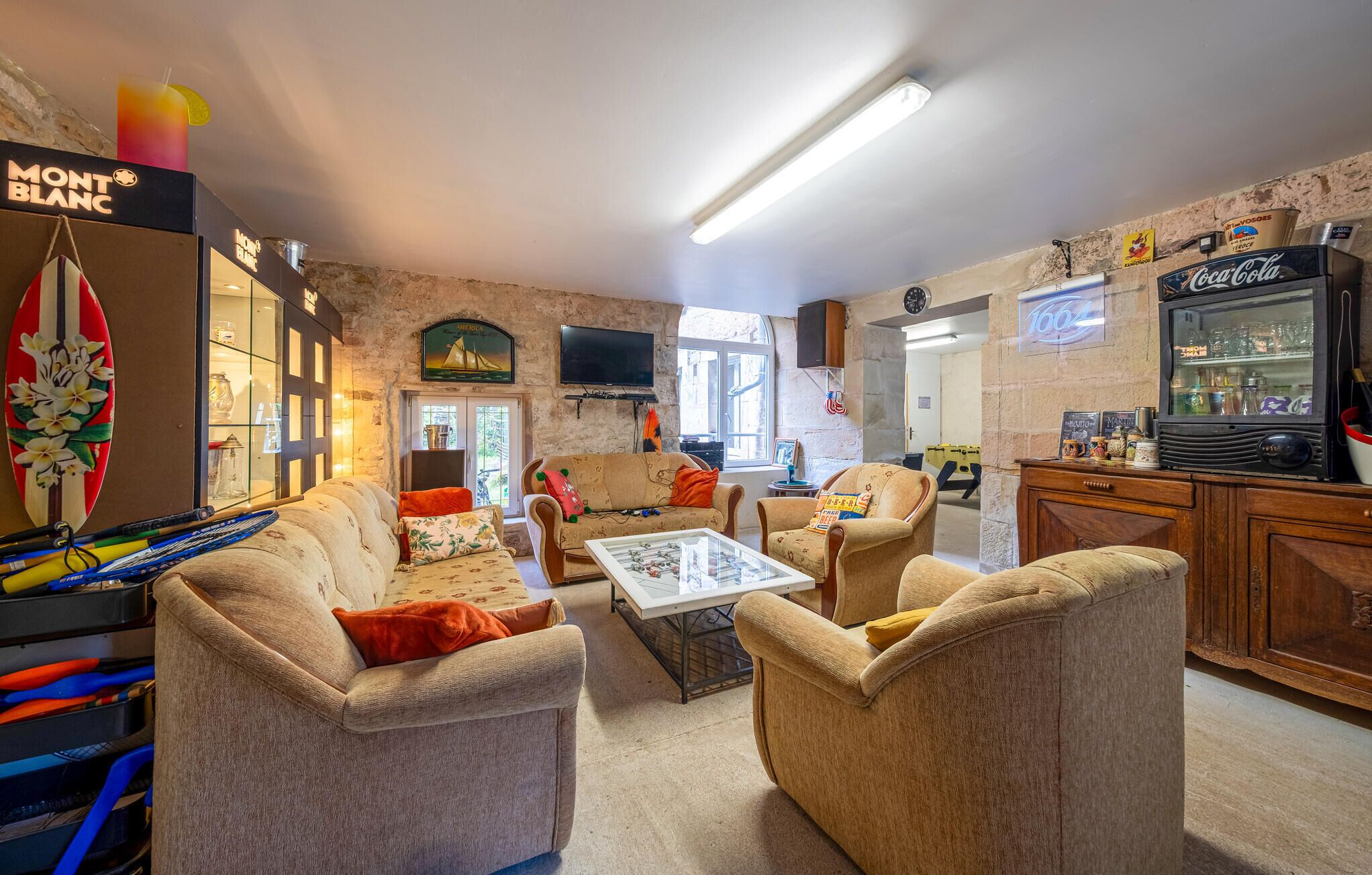 A cozy living room with a sofa, armchairs, a coffee table, and a television. There is a surfboard decoration, a Coca-Cola refrigerator, and a window with a view.