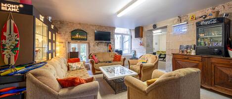 A cozy living room with a sofa, armchairs, a coffee table, and a television. There is a surfboard decoration, a Coca-Cola refrigerator, and a window with a view.