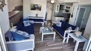 Un salon comprenant un canapé, une table basse et un téléviseur. On aperçoit une cuisine au fond de la pièce.