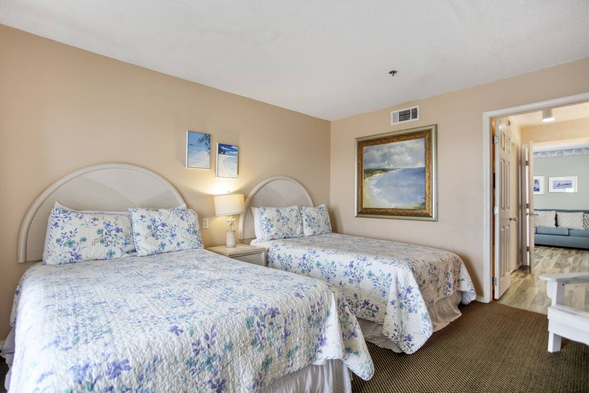 A bedroom with two beds, a nightstand, a lamp, a framed painting, and a view of another room.