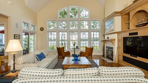 A spacious living room with a high ceiling, large windows, a fireplace, and a television. - Beautiful New Seabury Home in Mashpee (Mashpee)