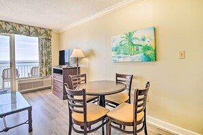 A dining area with a round table and four chairs, a TV, a lamp, and a painting of a tropical scene on the wall. - Oceanfront Myrtle Beach Condo w/ Balcony! (Myrtle Beach)