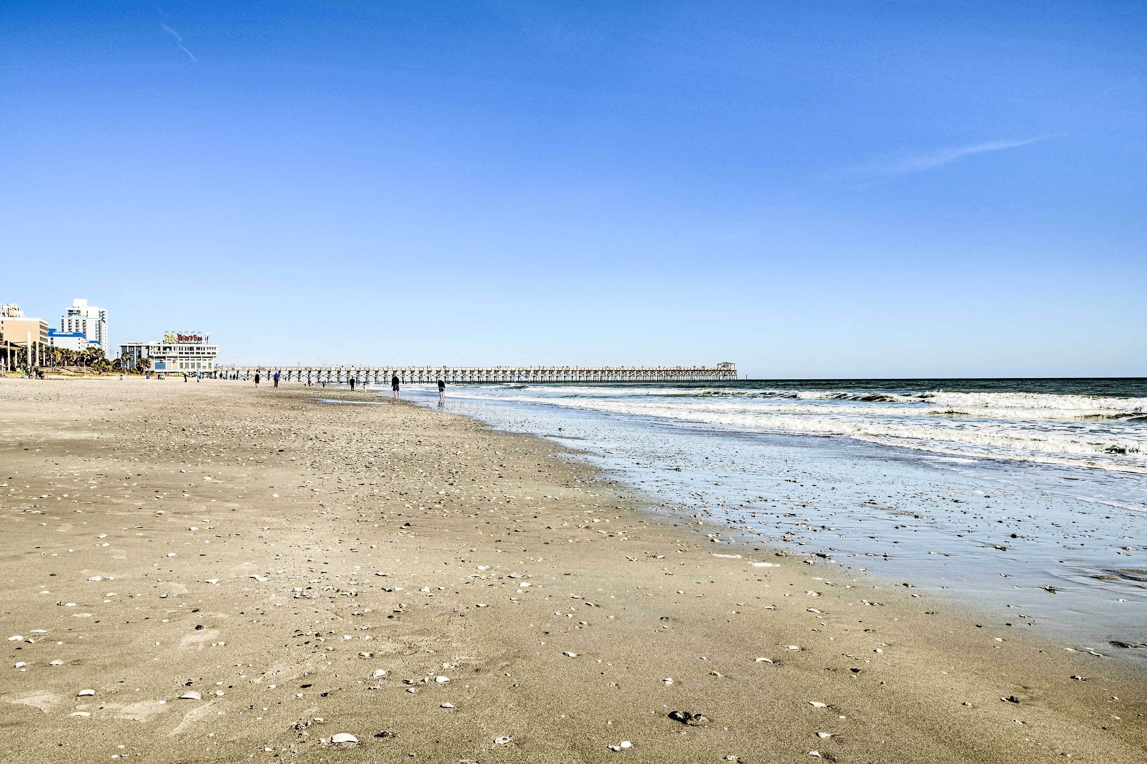 Una spiaggia con un molo che si protende sull'oceano, una riva sabbiosa e edifici sullo sfondo.