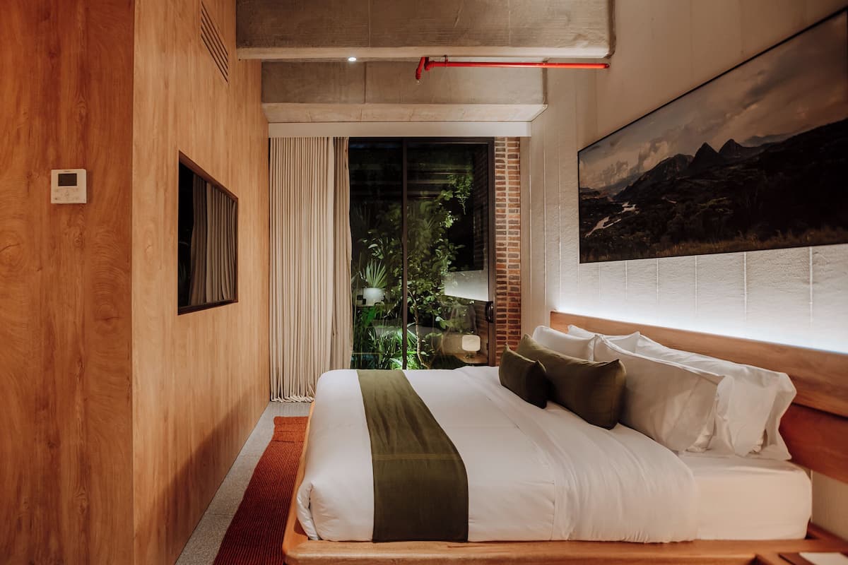 Habitación de hotel moderna con una cama grande, vistas a zonas verdes a través de un ventanal y un cuadro de paisaje montañoso en la pared.