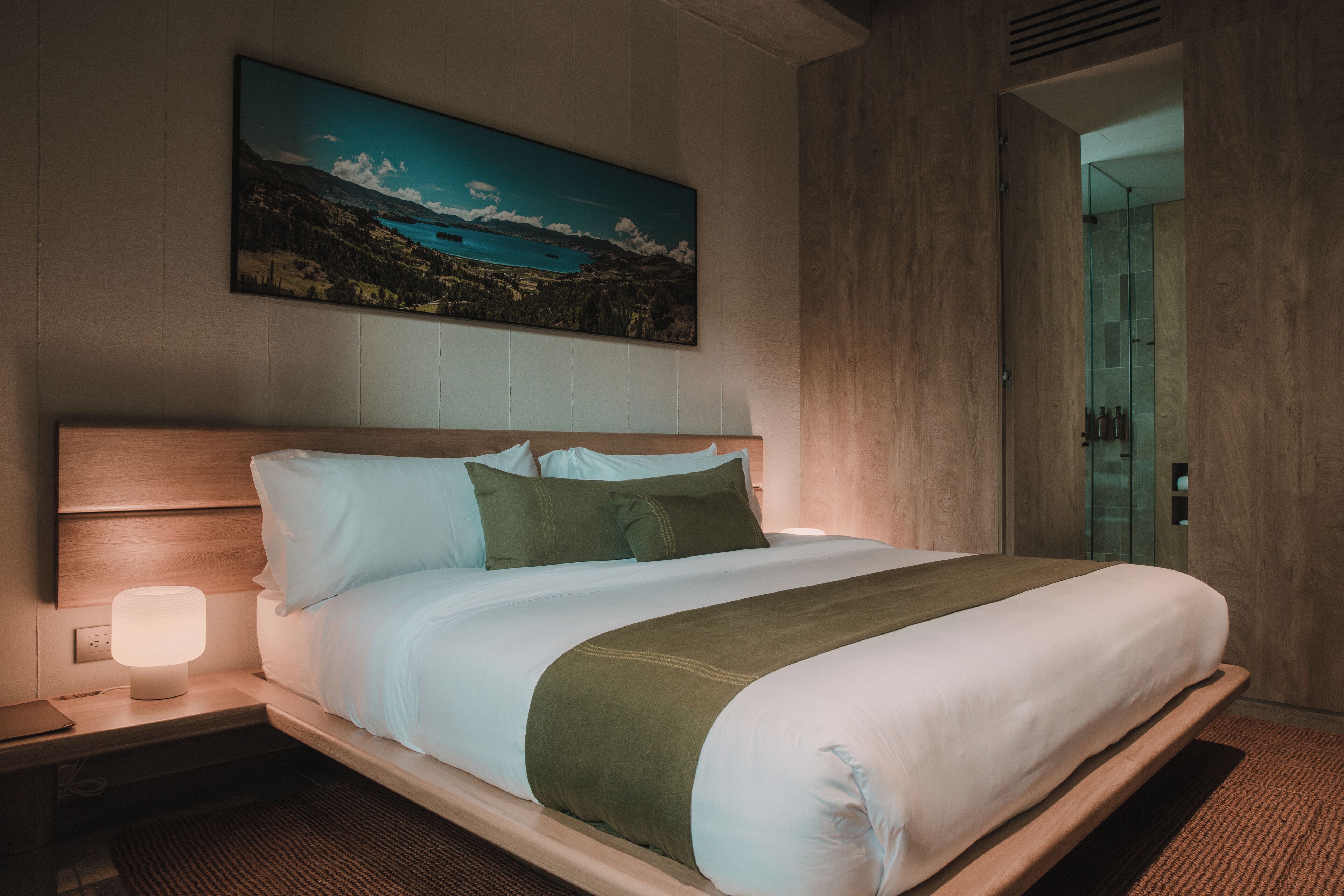 A neatly made bed with white and green bedding, a wooden headboard, and a nightstand with a lamp. A painting of a landscape is on the wall above the bed.