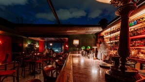 A bar with a long counter, chairs, and a bartender. There are shelves with bottles and a view of the city at night.