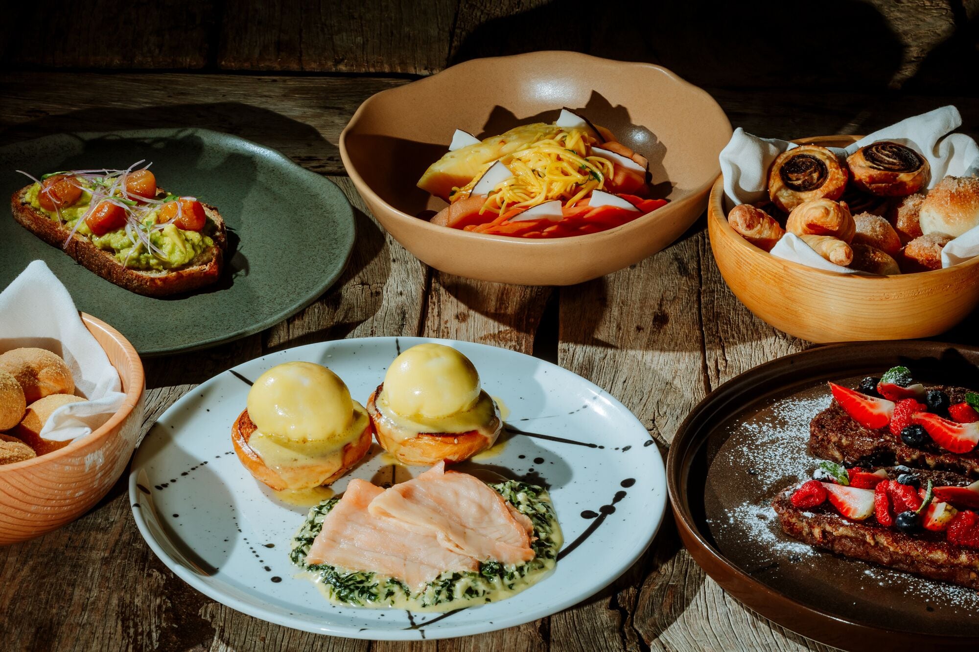 Verschiedene Speisen, darunter Eier Benedict, Lachs und Gebäck, auf einem rustikalen Holztisch.