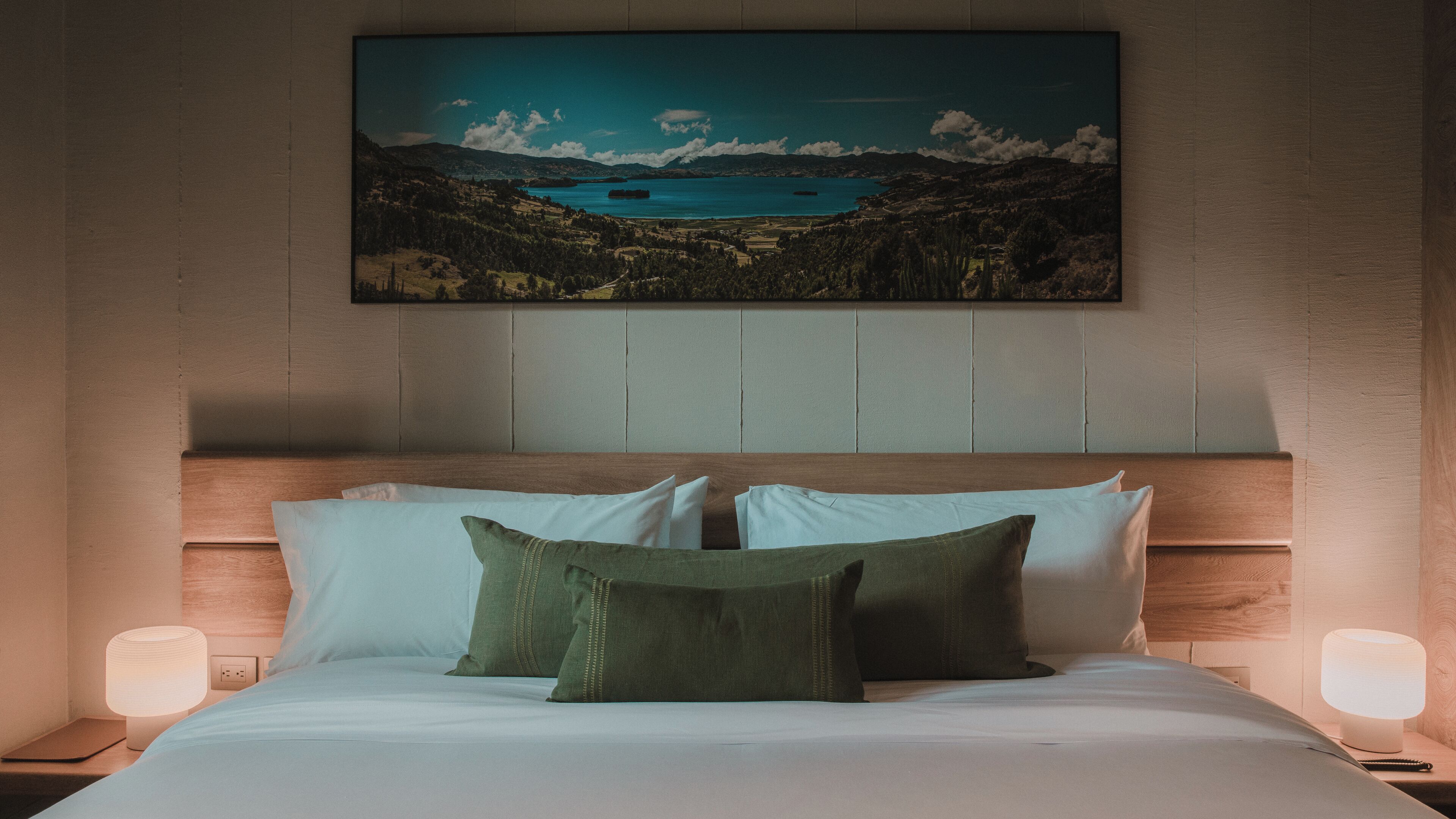 A bedroom with a large bed, two bedside lamps, and a painting of a landscape on the wall.