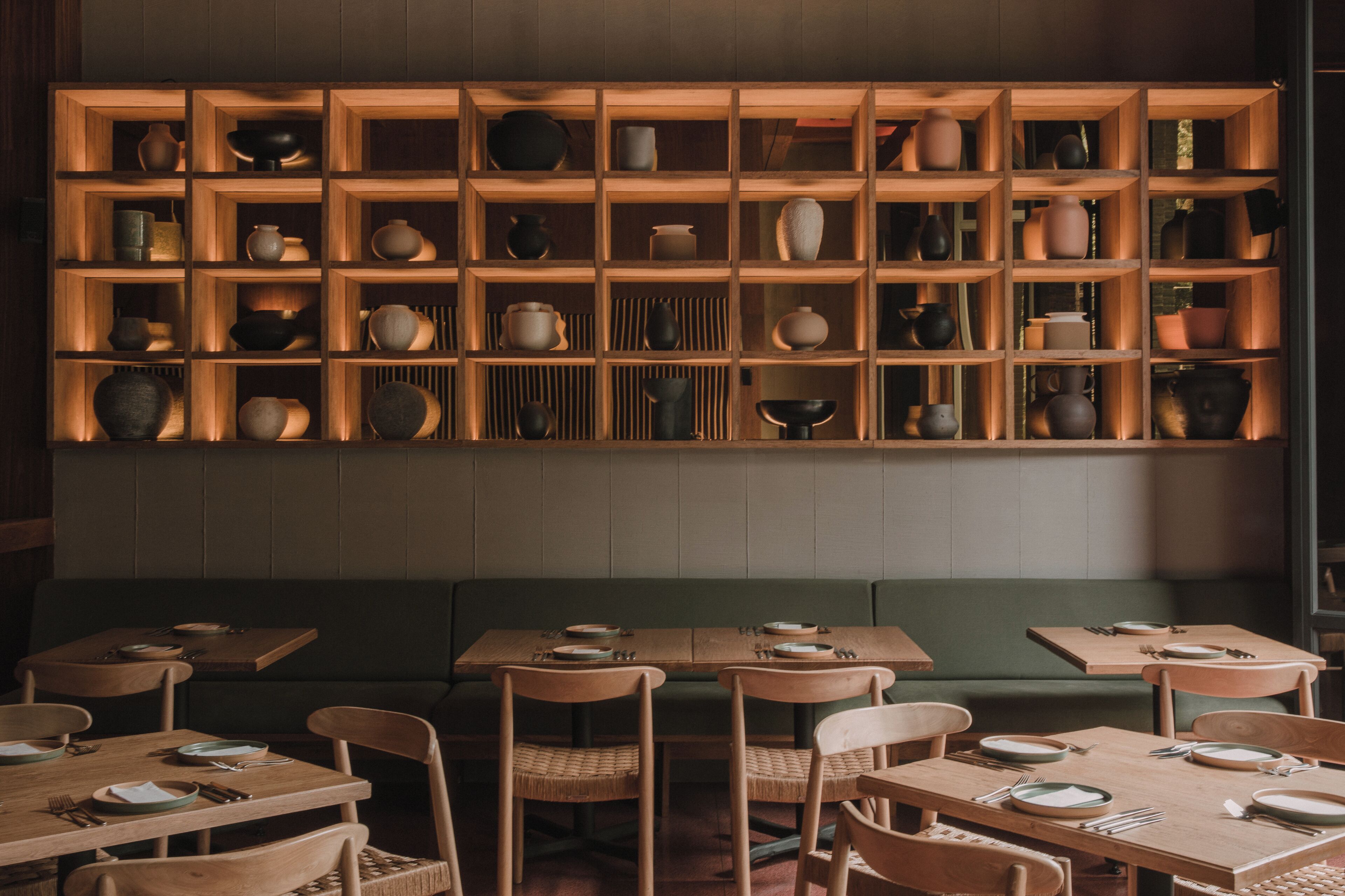 Un restaurant avec des tables et des chaises en bois, un coin salon vert et un mur recouvert d’étagères où sont exposées diverses pièces de poterie et de vaisselle.