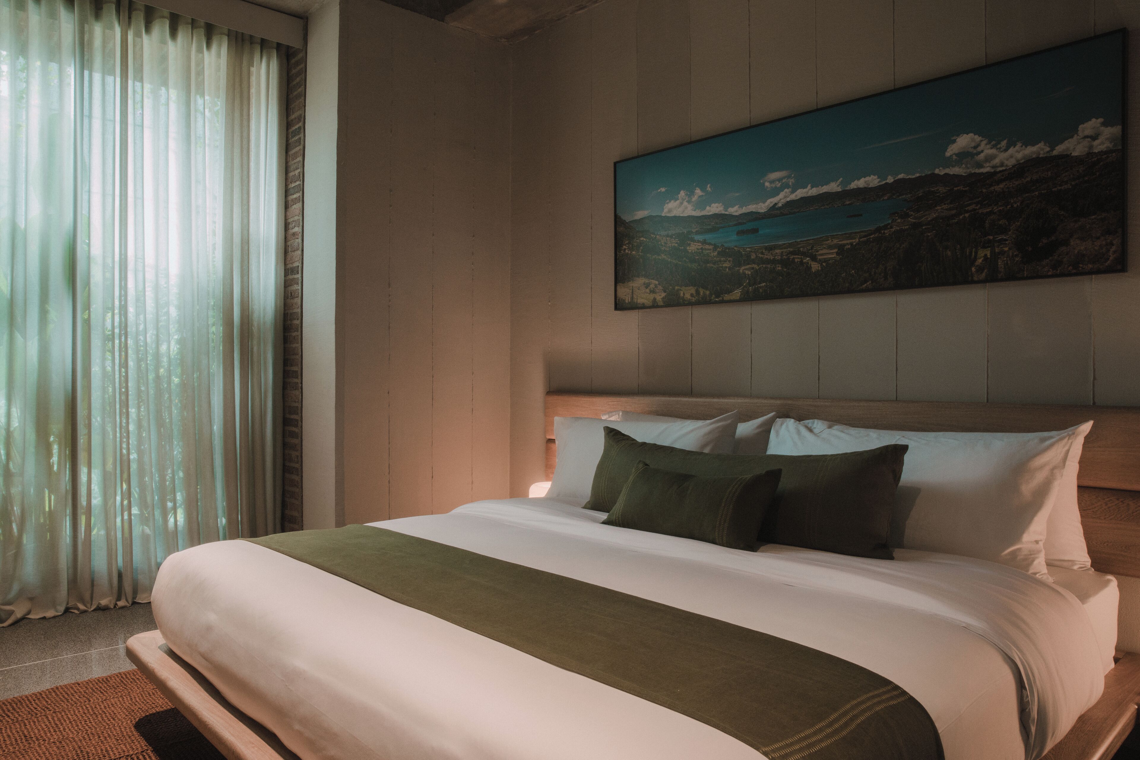 Una cama bien hecha con una manta verde, un cuadro de un paisaje y una ventana con cortinas traslúcidas.