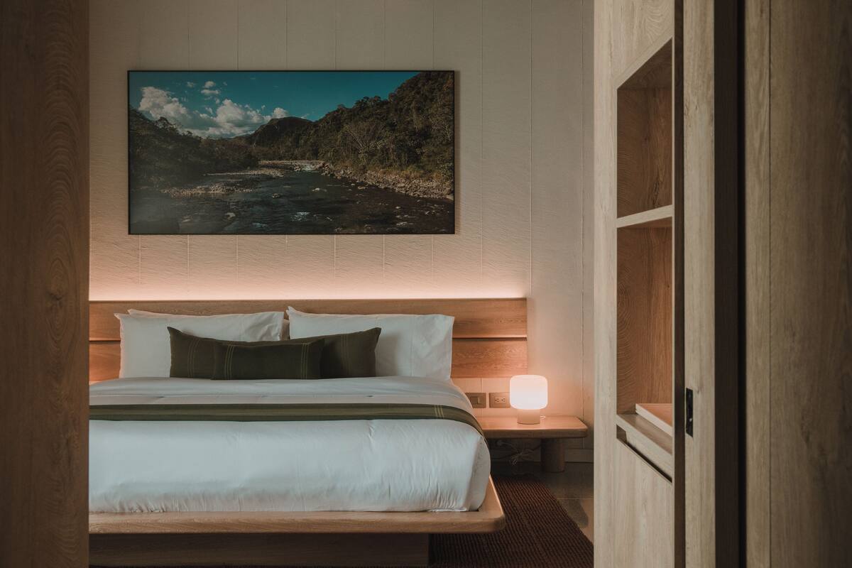 Un dormitorio con cabecera de madera, cama con ropa blanca, mesita de noche con lámpara y un cuadro de paisaje enmarcado sobre la cama.
