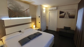 A hotel room with a large bed, a bedside table with a lamp, a sofa, and a wall-mounted TV. - Therra Lodge (Preganziol)