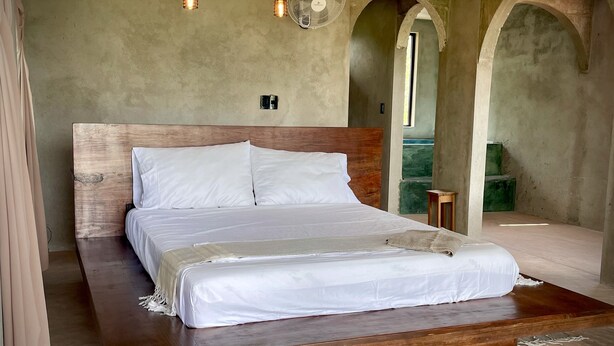A large bed with a wooden headboard, white bedding, and a wooden platform. There are two hanging light fixtures and a standing fan. The room has a rustic, concrete wall and an arched doorway. - CASA TÚA, amazing views just minutes to the beach! (Puerto Escondido)