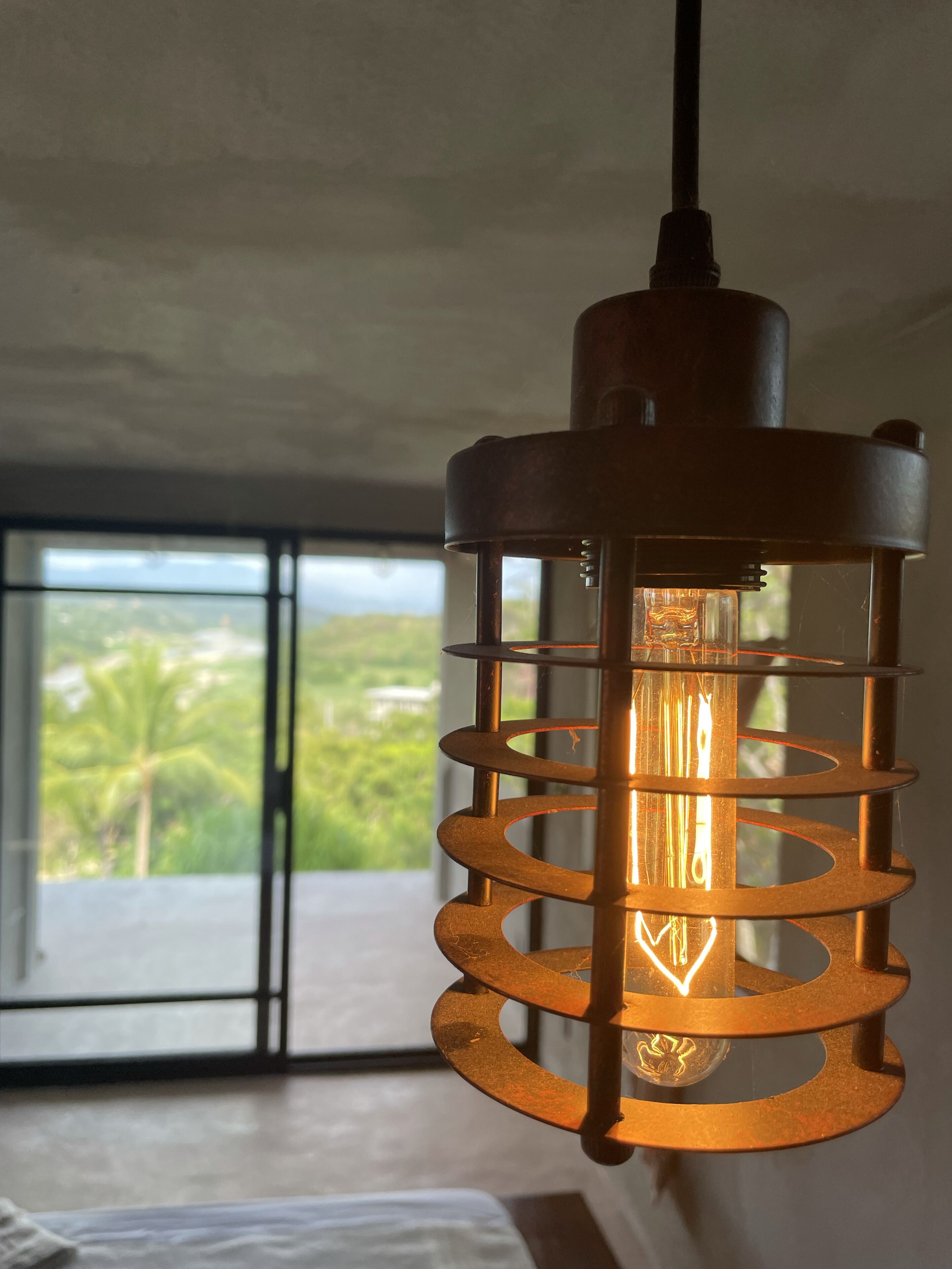A hanging light fixture with a spiral design, a visible bulb, and a view of a balcony with greenery outside.