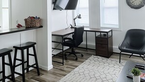 Un salon moderne avec un bureau, un fauteuil de bureau et un téléviseur fixé au mur. On y trouve deux tabourets de bar et une table basse agrémentée d’une plante.