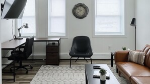 Un salon moderne comprenant un bureau, un fauteuil de bureau noir, un canapé en cuir marron, un tapis à motifs et une horloge murale.
