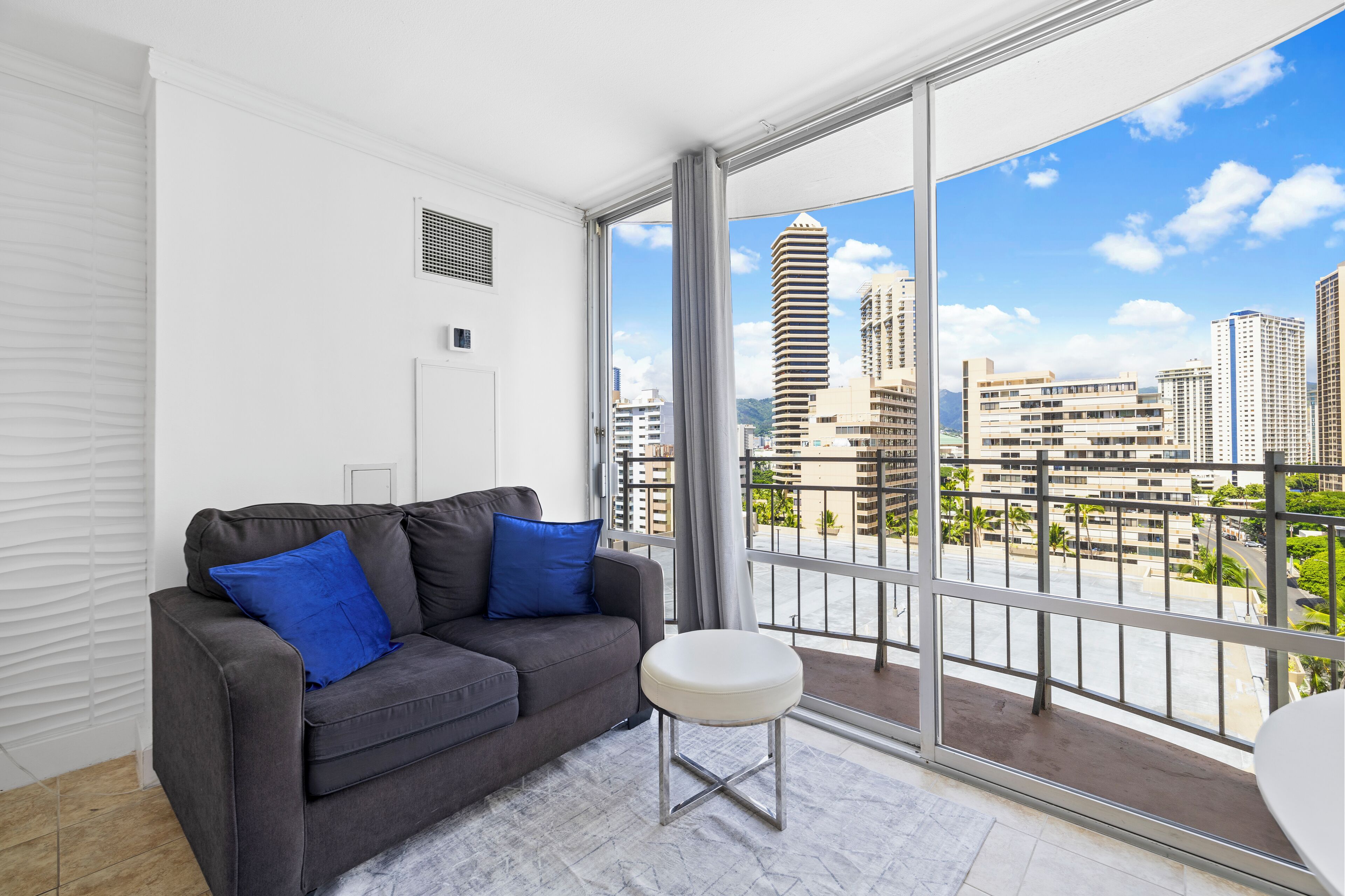 A modern living room with a grey sofa, blue cushions, a white ottoman, and a large window offering a city view.
