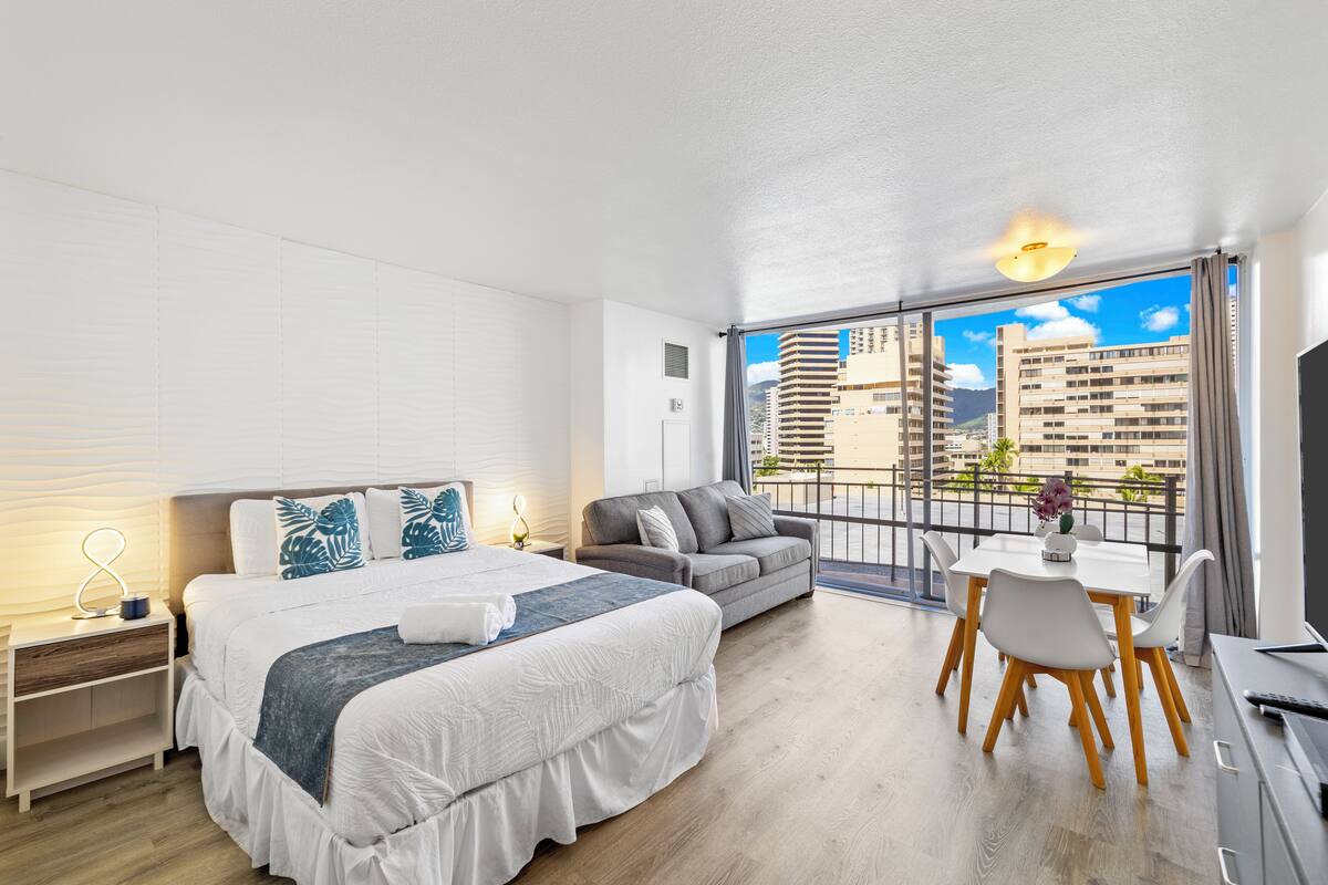 Quarto de hotel moderno com uma cama grande, um sofá, uma mesa de jantar e uma varanda com vista para a cidade.
