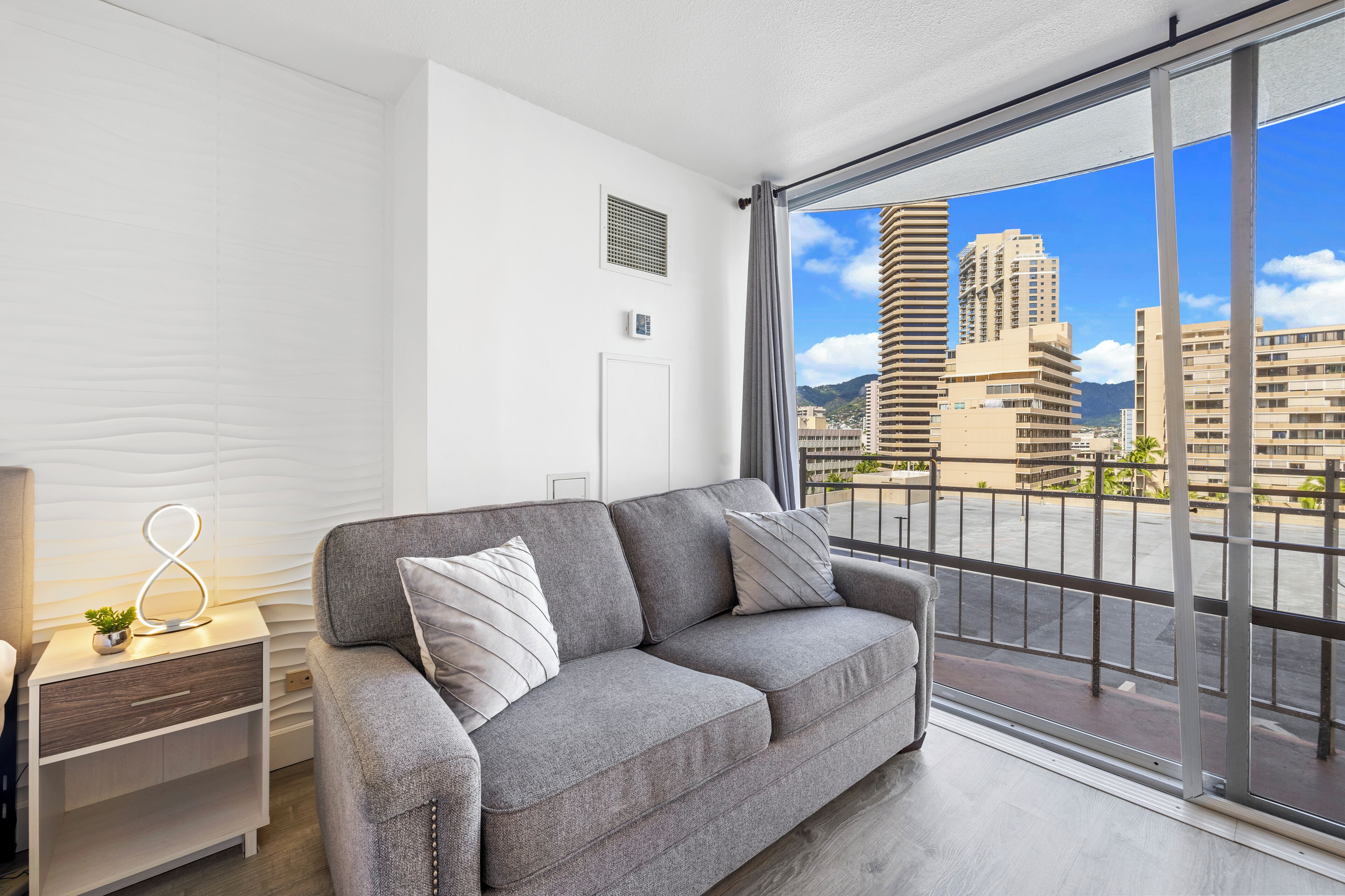 A modern living room with a grey sofa, a small side table with a plant, and a view of tall buildings through a large window.