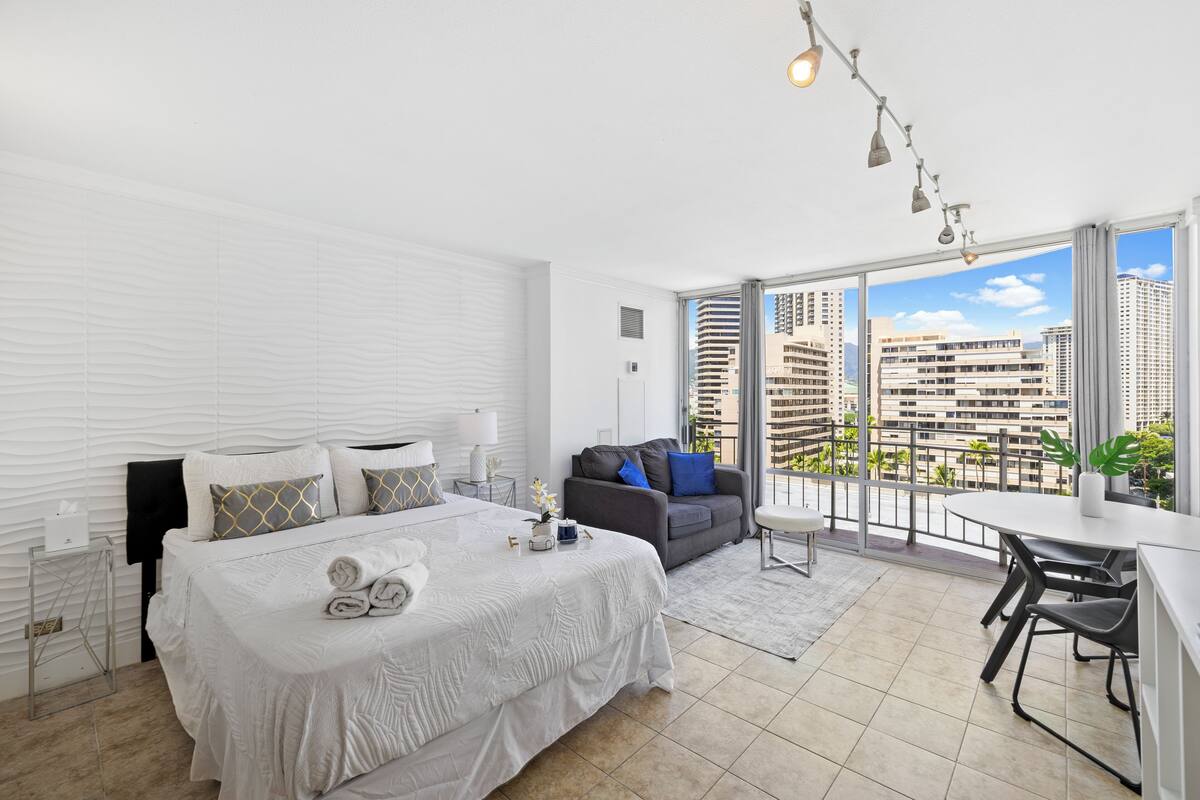 Quarto de hotel moderno com uma cama grande, um sofá, área de refeições e varanda com vista para a cidade.