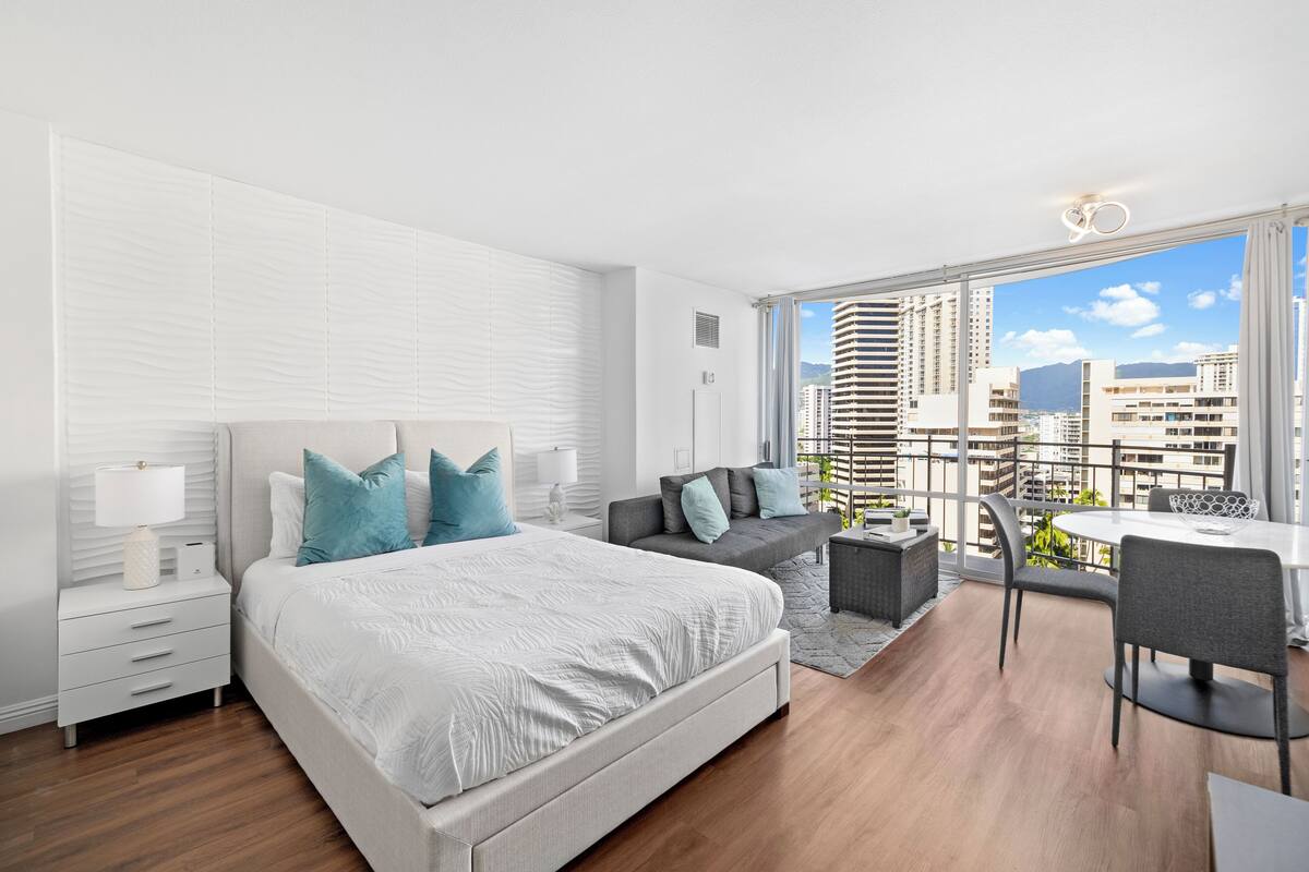 Quarto moderno com uma cama grande, um sofá cinza, uma mesa de jantar e vista para a cidade através de amplas janelas.