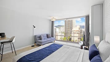 A modern hotel room with a large bed, a sofa, a desk, and a view of the city through large windows.
