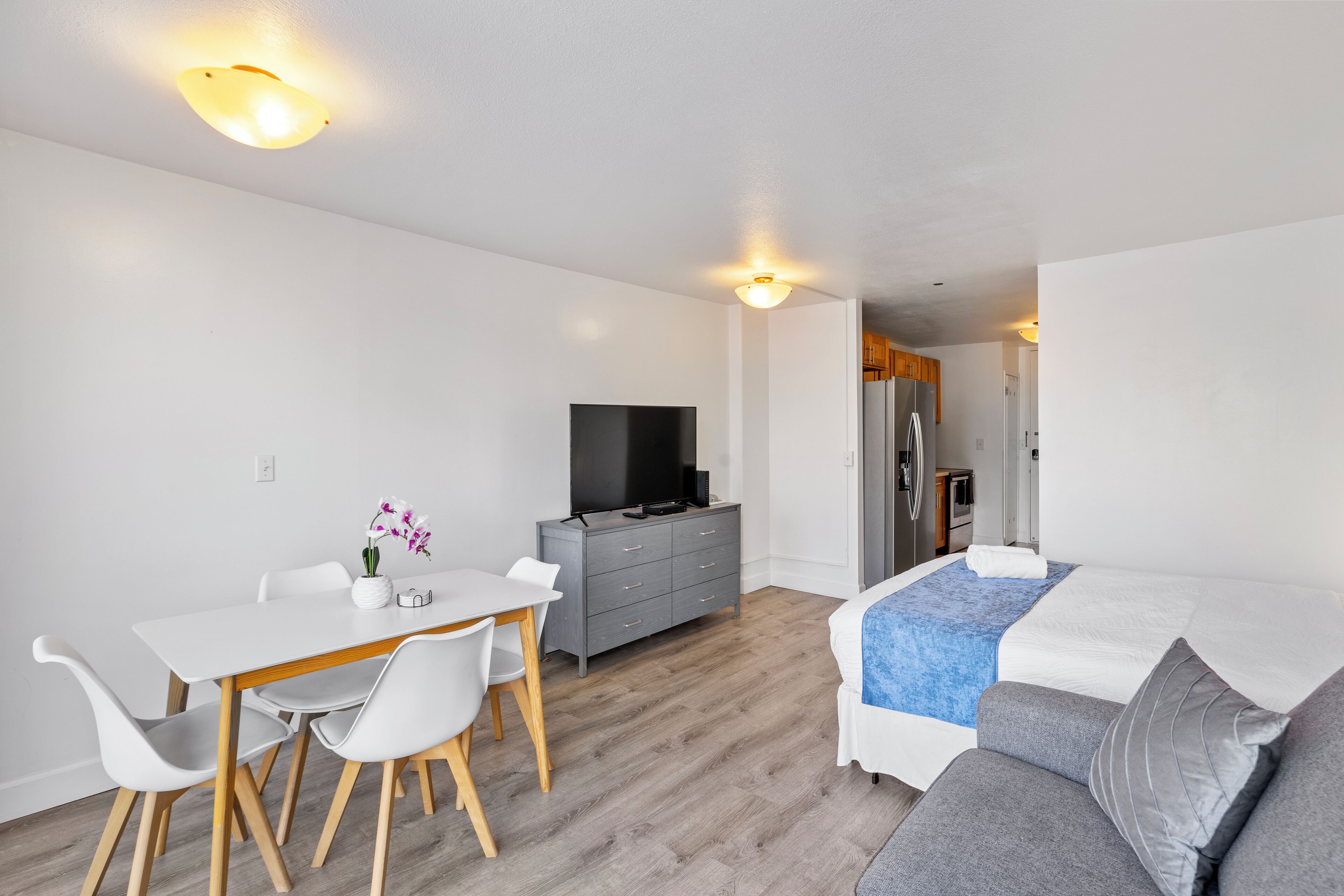 A modern, well-lit room with a dining table, chairs, a grey sofa, and a TV.