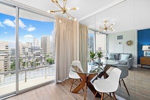 A modern living room with a glass dining table, white chairs, a grey sofa, and a city view through large windows.