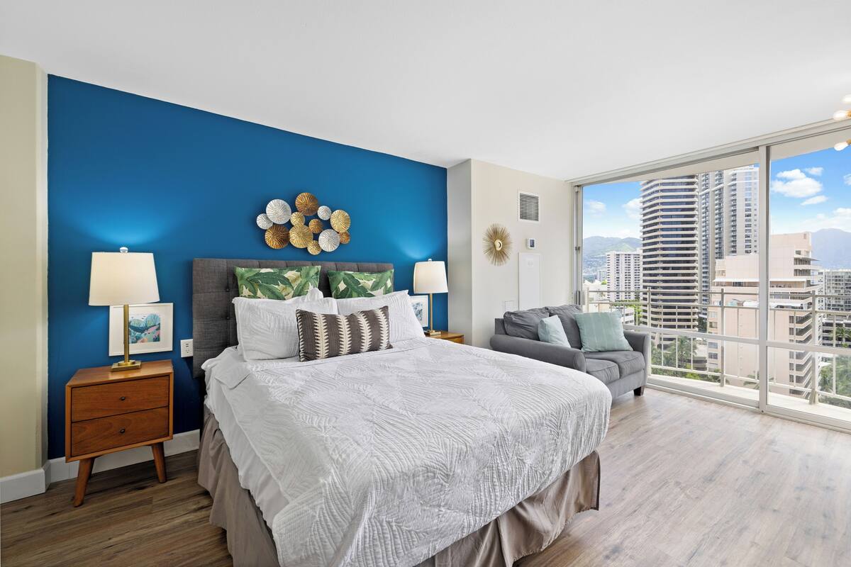 Quarto com uma cama grande, um sofá cinzento, uma mesa de cabeceira com abajur e vista para prédios altos através de grandes janelas.