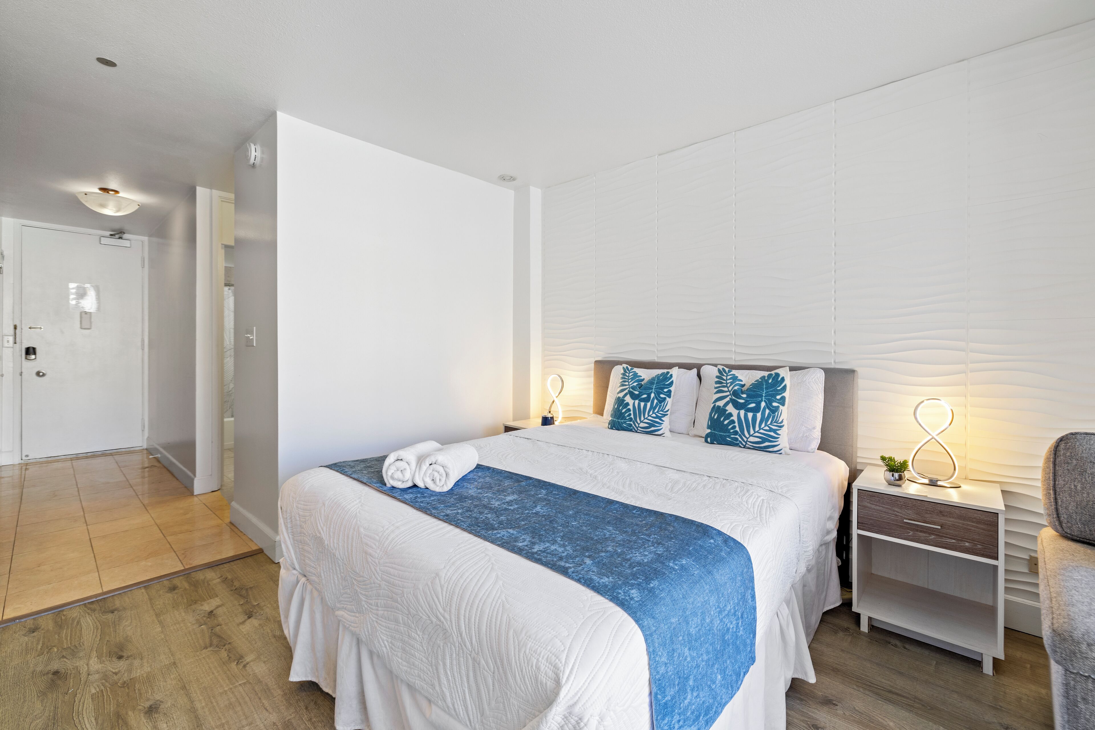 Quarto de hotel moderno com uma cama grande, uma mesa de cabeceira com abajur e vista para o corredor.