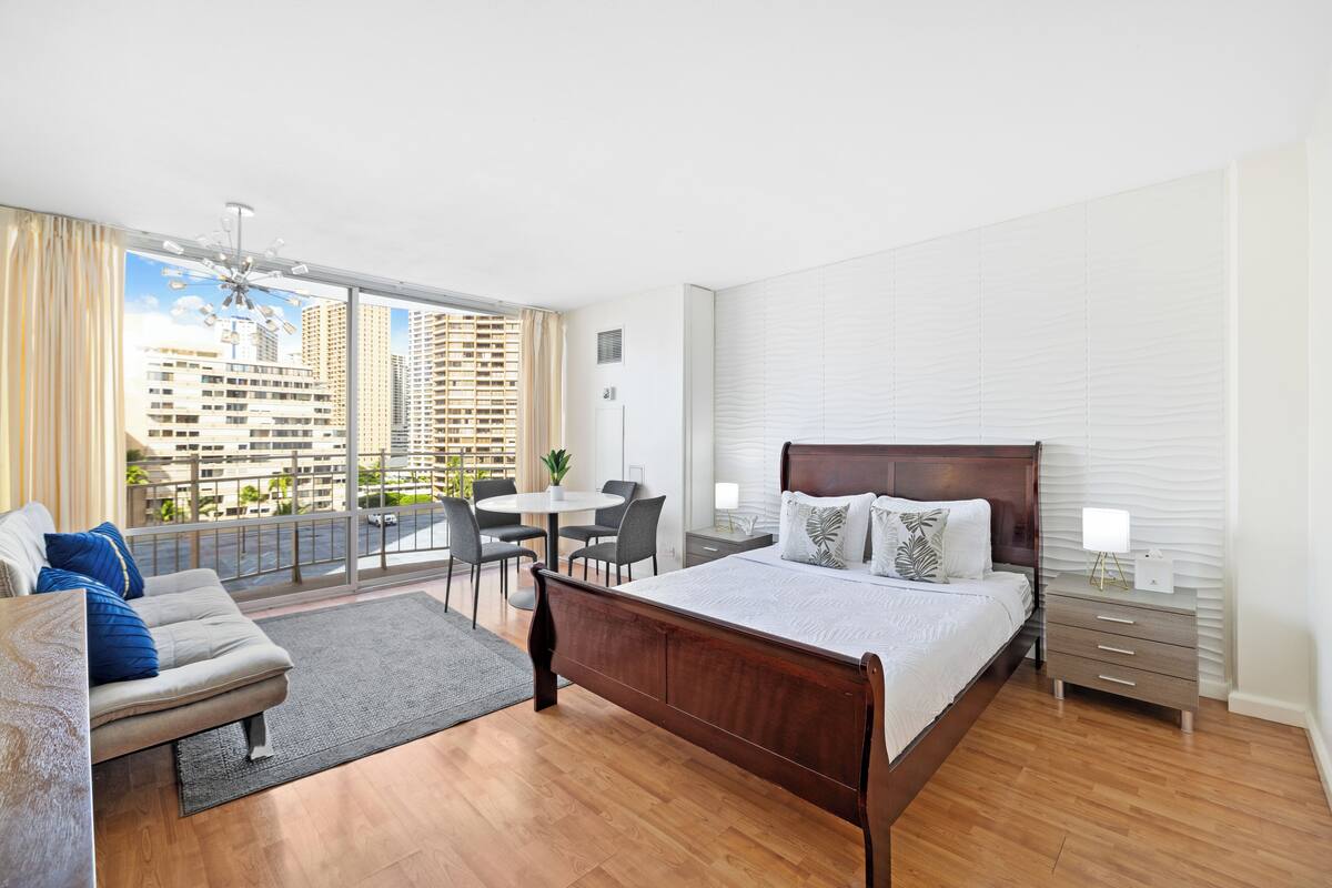 Quarto de hotel moderno com uma cama grande, um sofá, um espaço de refeições e uma varanda com vista para a cidade.