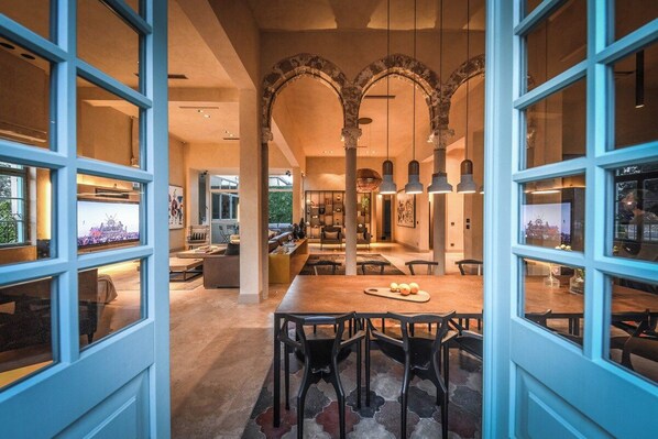 An open-plan living area with a dining table, chairs, and a view through large windows.