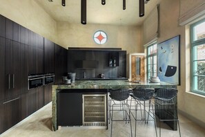 A modern kitchen with dark cabinetry, a marble countertop, and a built-in refrigerator. There are three black bar-style chairs and a large window with a view.
