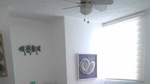 Ein Schlafzimmer mit Bett, Schreibtisch, Stuhl, Deckenventilator, Wandregal und Fenster mit Jalousien.