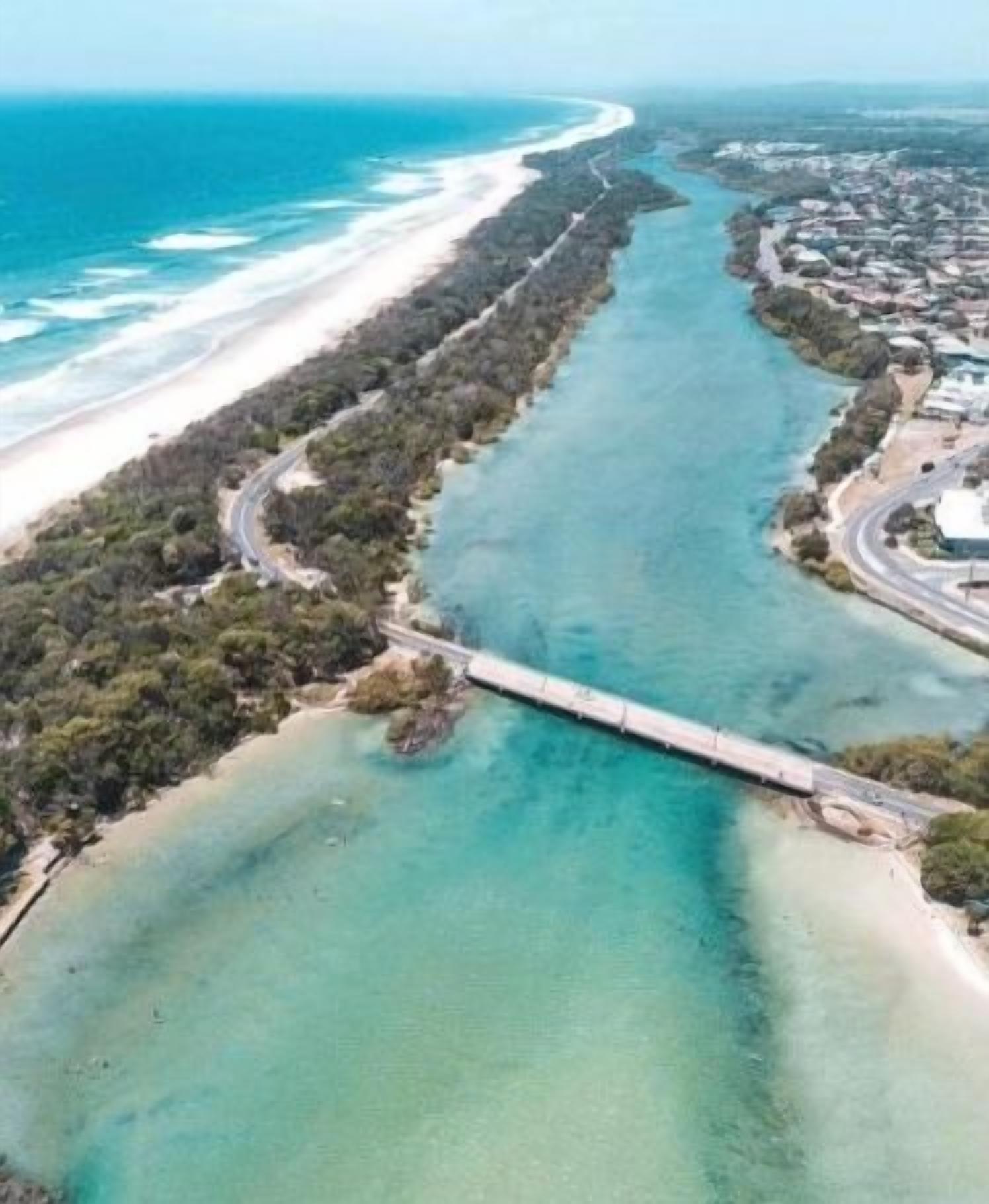 An aerial view of a coastal area with a long bridge connecting two land masses, clear turquoise waters, and a sandy beach.