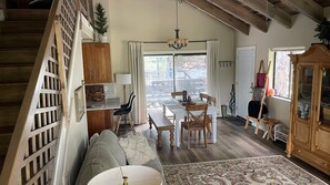 Un salon chaleureux avec un canapé, une table à manger et des chaises, un lustre, et une vue sur l’extérieur grâce à de grandes fenêtres.