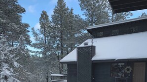 A modern cabin with a dark exterior, a covered porch, and a snow-covered landscape. - Mountain Pine Main House on Quiet Cul-de-sac-3 Decks-Views of Forest + Hot Tub! (Flagstaff)