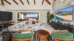 A spacious living room with a large flat-screen TV, a sectional sofa, and a dining area with a view of the beach.