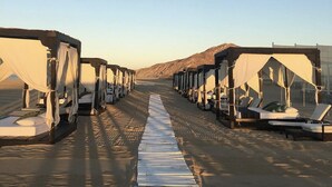 A row of tents with beds and lounge chairs, arranged on a sandy path with a clear sky and mountains in the background.