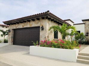 A house with a stone facade, a dark garage door, and a small garden with pink flowers. - Luxury Panoramic Ocean View Private Pool @Coronado (Cabo San Lucas)