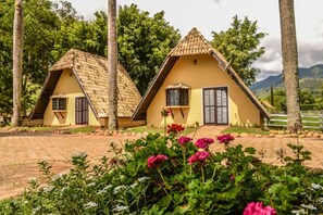 Two thatched-roof cottages with a paved pathway and lush greenery in the background. - VILLA DAS PALMEIRAS Empório & Chalés (São Bento do Sapucaí)