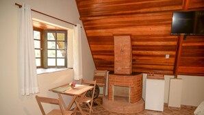 A cozy room with a wooden ceiling, a brick fireplace, a small table with chairs, a window with curtains, and a television mounted on the wall. - VILLA DAS PALMEIRAS Empório & Chalés (São Bento do Sapucaí)