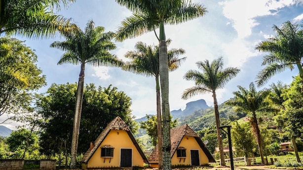 A row of yellow cottages with thatched roofs, surrounded by lush greenery and palm trees, set against a backdrop of mountains. - VILLA DAS PALMEIRAS Empório & Chalés (São Bento do Sapucaí)