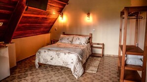 A bedroom with a sloped ceiling, a bed with floral bedding, a wooden nightstand, a bench, and a wall-mounted shelf. - VILLA DAS PALMEIRAS Empório & Chalés (São Bento do Sapucaí)