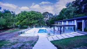 A modern outdoor pool area with a clear view of the pool, surrounded by lush greenery and a covered patio with seating. - Southern Highlands Finest! (Exeter)