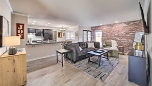 A modern living room with a grey sofa, a coffee table, and a TV on a stand. There is a kitchen in the background with a brick wall and a dining area with a table and chairs. - Immaculate 'Woodland View' Retreat w/ Hot Tub (Tulsa)