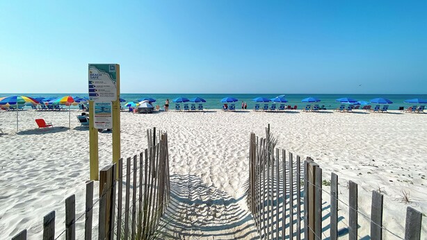 Ein Strand mit einer Holzbank, die zum Wasser führt, blauen Sonnenschirmen und Liegestühlen.